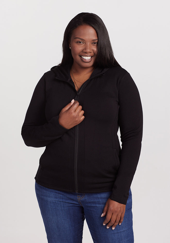 A person with long dark hair smiles while wearing the Woolx Zoey Zip Up Hoodie - Black and blue jeans. They are standing against a plain, light-colored background. | Le'Quita is 5'11", wearing a size XL