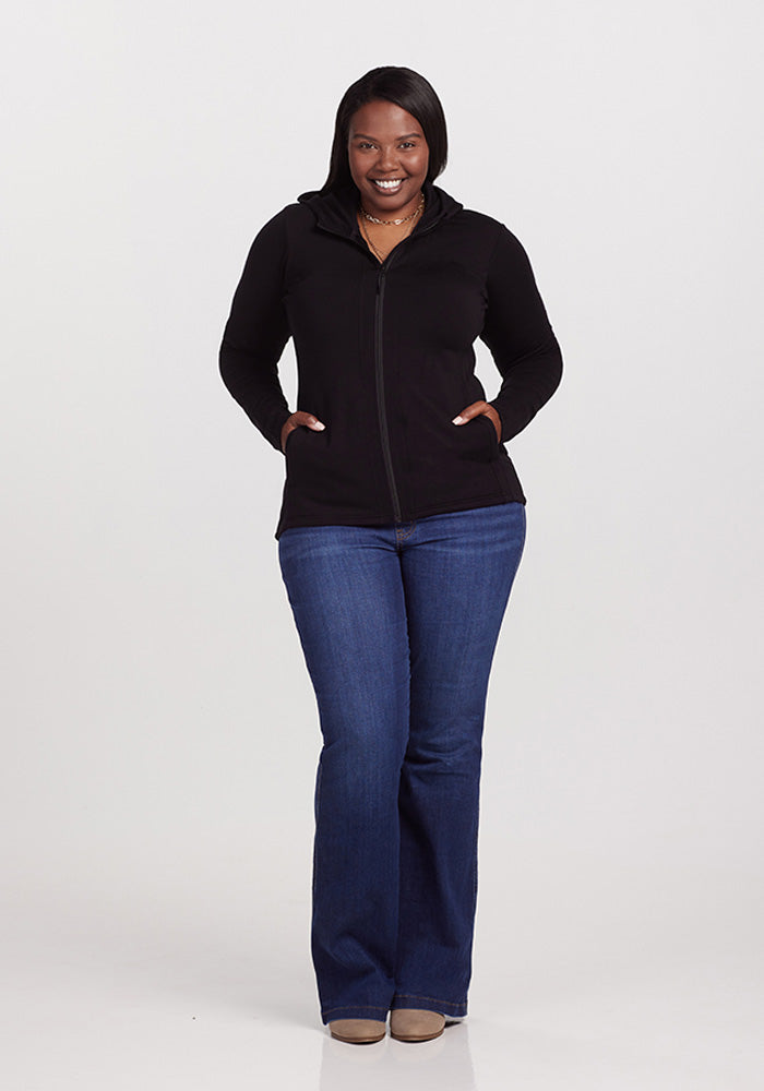 A woman with shoulder-length dark hair stands smiling, wearing the Woolx Zoey Zip Up Hoodie in black, blue jeans, and beige shoes. She has her hands in the pockets of this warm hoodie. The background is plain and white.