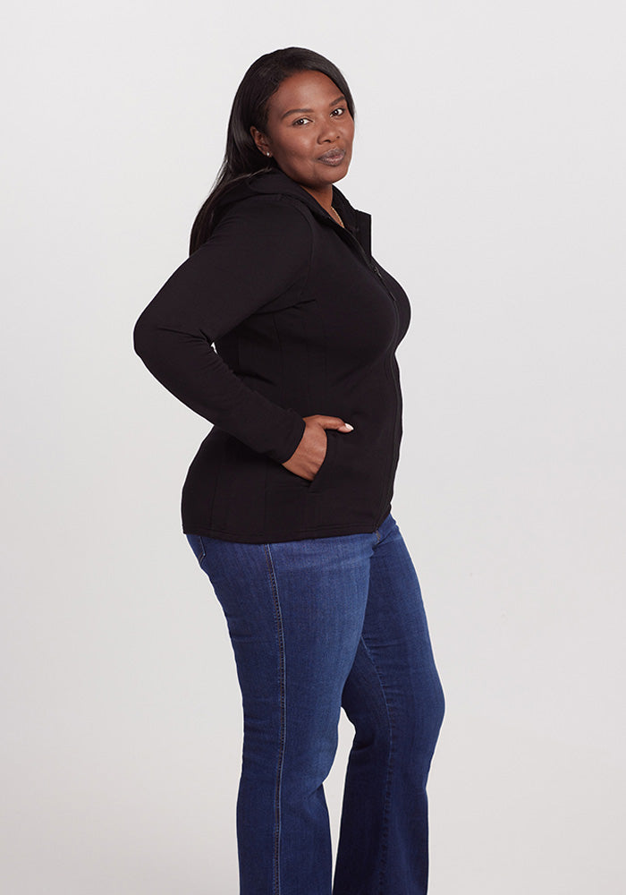 A person with long dark hair wearing a Woolx Zoey Zip Up Hoodie - Black, made from Woolx DuraLite® Fabric, and blue jeans stands against a plain white background. They have one hand in the hoodie pocket and are looking towards the camera with a neutral expression.