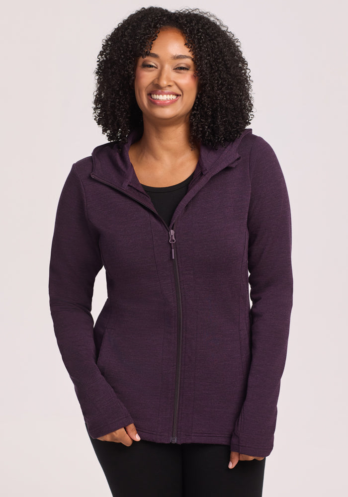 A woman with curly hair smiles wearing the Woolx Zoey Zip Up Hoodie • Final Sale in Deep Plum over a black top, standing against a plain light background.