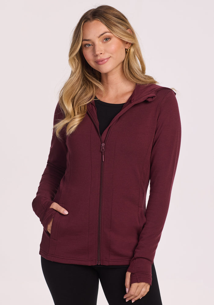 A woman with long blonde hair wears the Woolx Zoey Zip Up Hoodie in Toasted Garnet over a black top, standing with one hand in her pocket and smiling softly against a plain light background.
